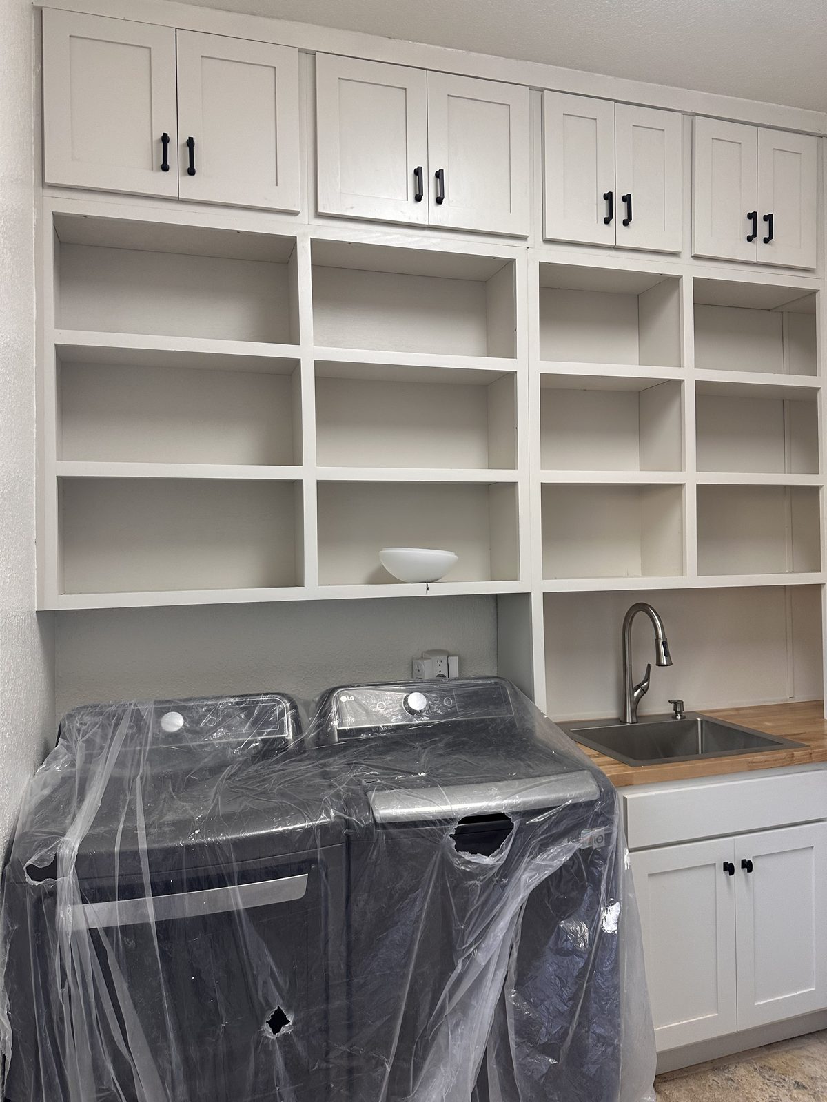 Custom white shaker laundry room cabinets with open cubby shelves above the washer and dryer and a sink set in a butcher-block countertop